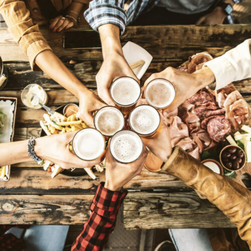 Friends Cheering Beer Glasses On Wooden Table Covered With Delicious Food - Top View Of People Having Dinner Party At Bar Restaurant - Food And Beverage Lifestyle Concept